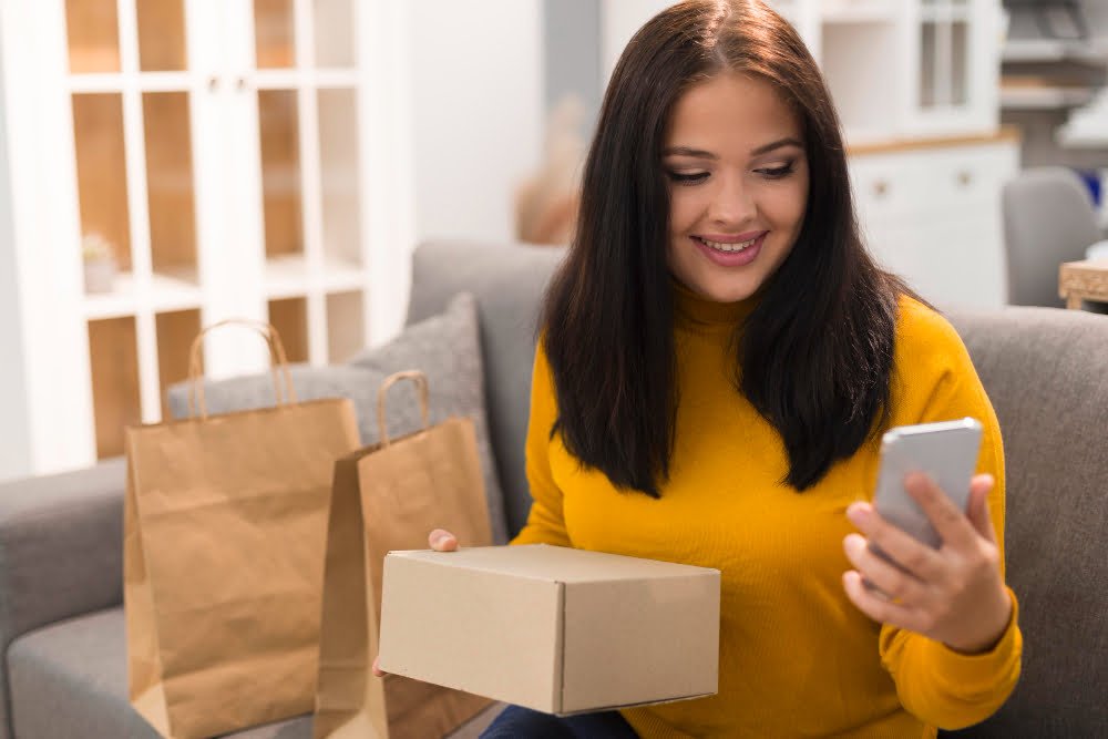 Mulher sentada em um sofá, segurando uma caixa parda, típica de envio de encomendas online. Na outra mão, um aparelho celular e, ao seu lado, algumas sacolas, também de envio de pedidos realizados via e-commerce.