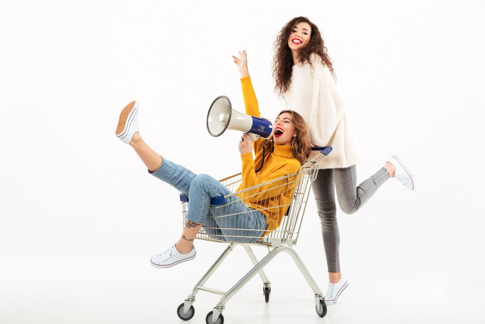 Duas mulheres: uma delas esta dentro de um carrinho de supermercado, com um mega fone, enquanto a outra a empurra, simulando a divulgação de uma marca, produto ou serviço